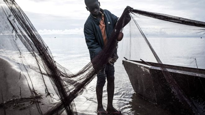 A young Congolese fisherman tends to his nets along the banks of Lake Tanganyika in Kalemie, Democratic Republic of the Congo. His trousers are rolled up and he is wearing a polo shirt with a quilted jacket over it.