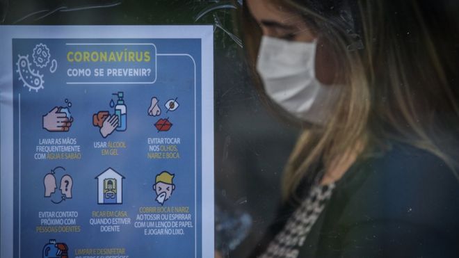 Mulher com máscara aparece de perfil e ao lado de cartaz intitulado 'Coronavírus: como se prevenir?'