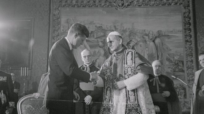 Черно-белая фотография, 1963 г., показывает, что Дж.Ф.К. пожимает руку Папе, в окружении многочисленных высокопоставленных священнослужителей среди пышной обстановки в Ватикане