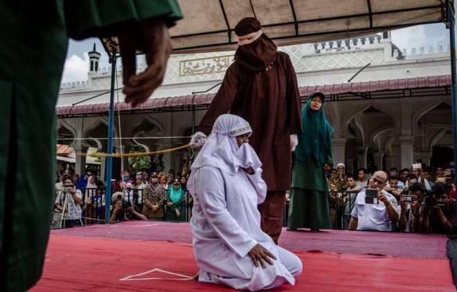 A woman on her knees is flogged in Aceh state by a hooded man