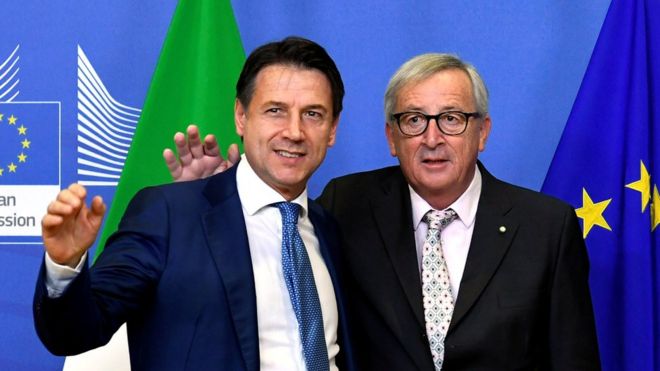 Джузеппе Конте машет в камеру, в то время как Жан-Клод Юнкер, кажется, собирается обнять итальянского премьер-министра в окружении итальянских и европейских флагов
