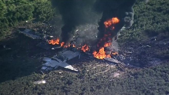 A still image showing a crashed plane in flames in a field, taken from video footage