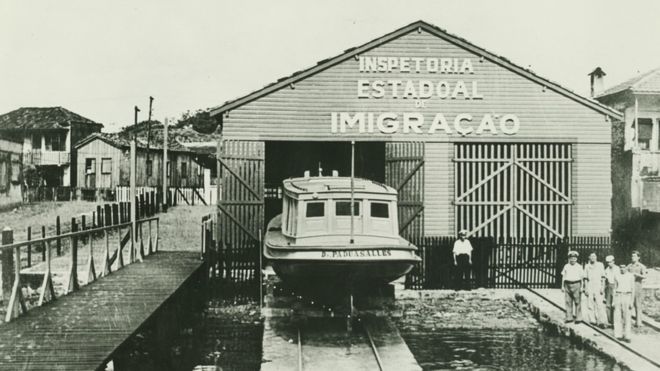Foto em preto e branco mostra pavilho, do qual sai barco e cuja fachada diz: Inspetoria Estadoal de Imigrao