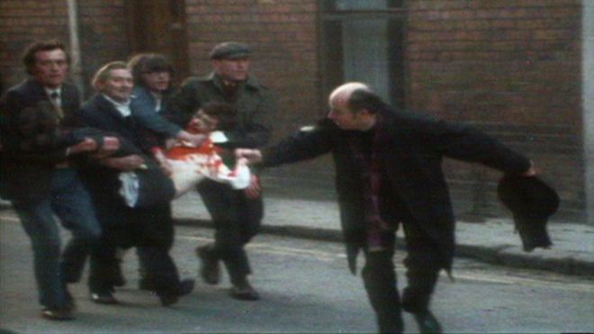 Священник машет окровавленным платком, пока четверо мужчин несут раненого, окровавленного человека по улицам Лондондерри в Кровавое воскресенье