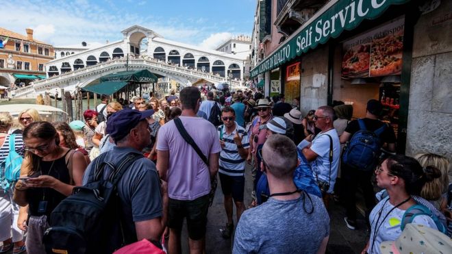 Большое количество людей толпится на набережной в Венеции, а на заднем плане виден знаменитый туристический район - мост Риальто