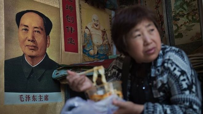 Продавец ест лапшу рядом с плакатом покойного китайского председателя Мао Цзэдуна (слева) на рынке в Пекине 15 мая 2016 года.