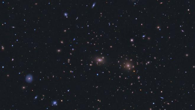 Звездное поле с множеством видимых галактик, как синих, так и белых. Они несколько сгруппированы ближе к центру изображения.