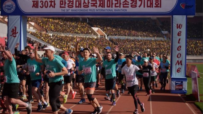Международные бегуны пересекают стартовую линию марафона в Пхеньяне, машут толпе на стадионе имени Ким II Сена