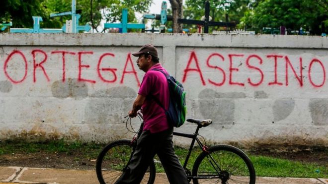 Un hombre camina delante de u muro que dice "Ortega asesino"
