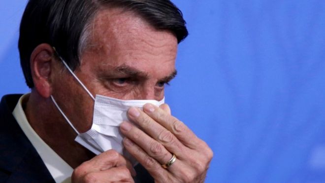 Em frente a painel azul, Bolsonaro aparece de lado ajeitando máscara