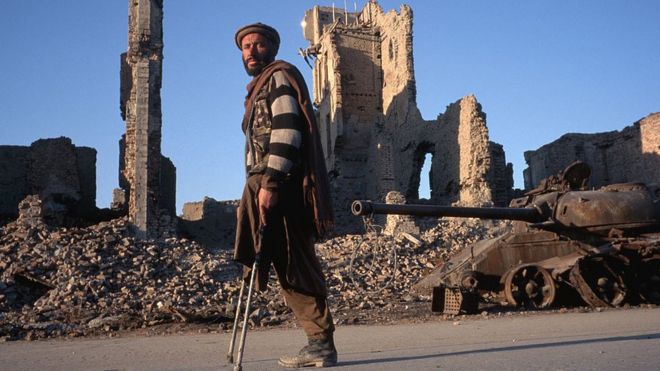 Мужчина, потерявший ногу на фугасе, идет мимо разрушенных бомбами зданий