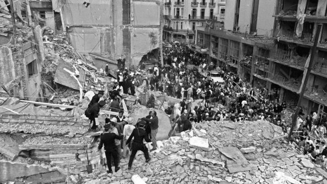 Пожарные, полицейские и спасатели разыскивают в руинах, а толпа наблюдает за тем, как 18 июля 1994 года в израильско-аргентинской ассоциации взаимопонимания (Amia) в Буэнос-Айресе взорвалась бомба,