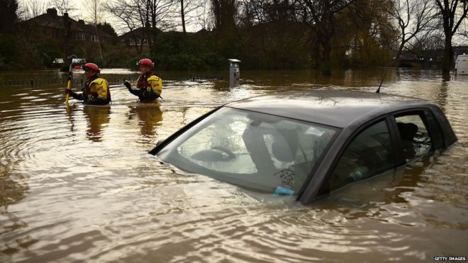 Автомобиль окружен и почти покрыт паводковой водой. Два спасателя позади, пробираются по воде.