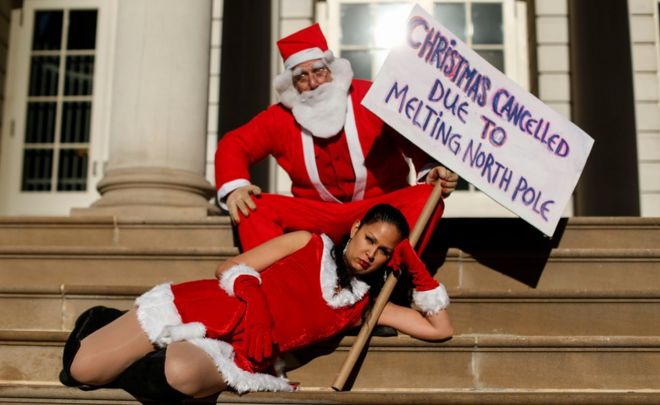 Активисты наряжаются, поскольку Санта-Клаус держит знамя, когда он принимает участие в глобальном климатическом марше в мэрии 29 ноября 2015 года в Нью-Йорке.