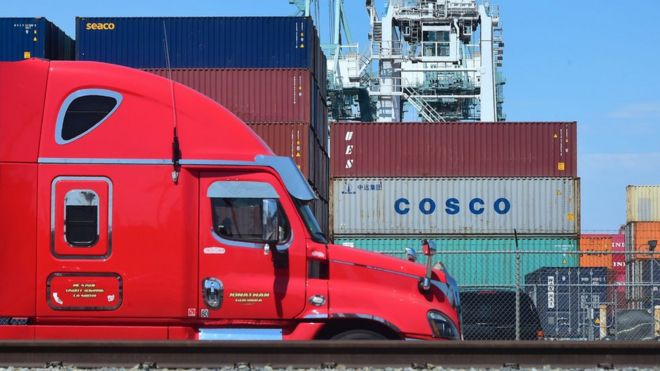 На этой фотографии, сделанной 6 июля 2018 года, грузовик для доставки контейнеров пропускает контейнеры, штабелированные в порту Лонг-Бич в Лонг-Бич, штат Калифорния, в том числе из COSCO, китайской государственной судоходной и логистической компании.