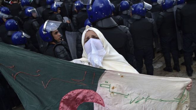 Mwanamke wa Algeria akiwa ameshikilia bendera ya taifa karibu na vikosi vya usalama vilivyozingira waandamanaji wakati wa maandamano ya kupinga mfumo wa utawala nchini humo katika mji mkuu wa Algeria, Algiers Jumatano tarehe 10 Aprili 2019