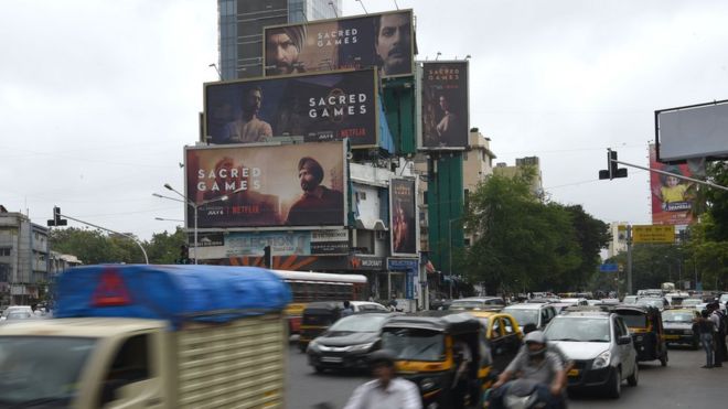 На этом снимке, сделанном 4 июля 2018 года, изображены индийские пассажиры, проезжающие мимо больших рекламных щитов для предстоящей индийской серии «Священные игры» на Netflix в Мумбаи.
