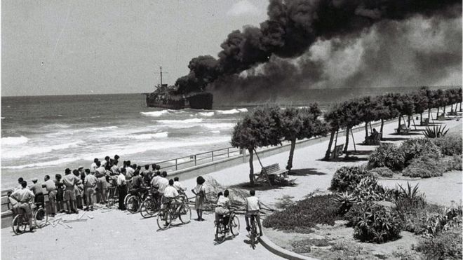 Navio pegando fogo e povo observando na praia