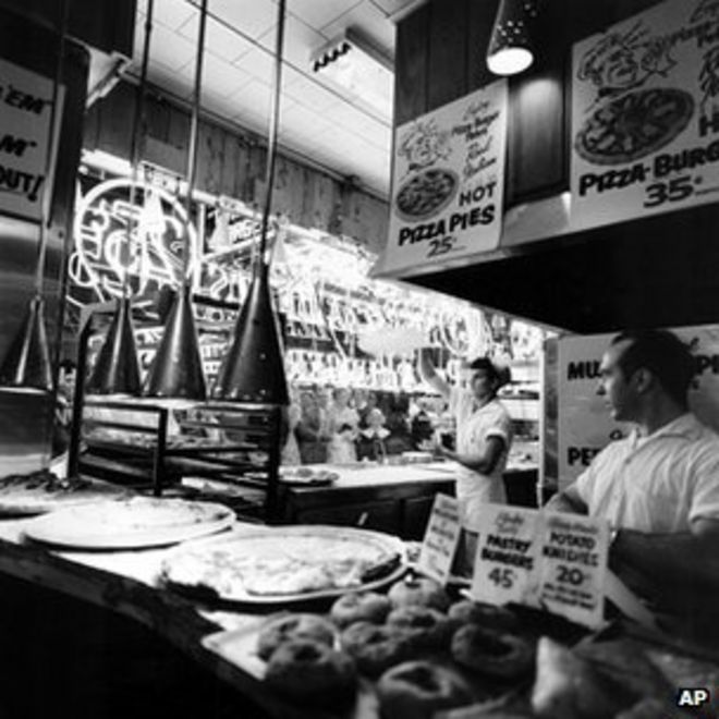 Прохожие наблюдают, как мужчина раскручивает тесто в воздухе, чтобы сделать пирог с пиццей в пиццерии на Таймс-сквер в Нью-Йорке, суббота, 22 февраля 1958 года.