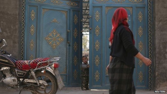 Подайте фотографию ребенка, выглядывающего из двери, когда мимо проходит уйгурская женщина в жилом районе в Турфане, Синьцзян