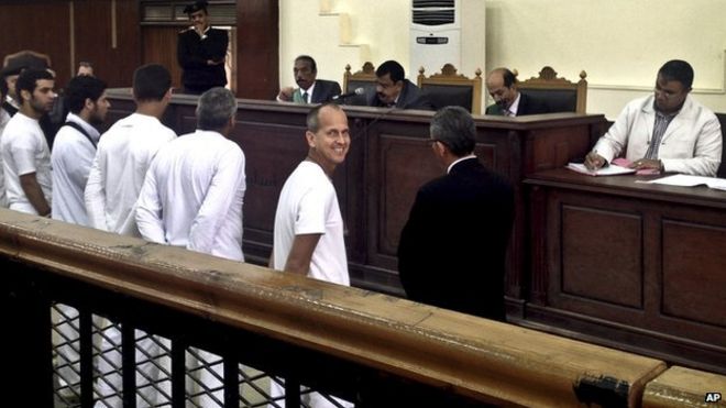 Английский корреспондент «Аль-Джазиры» Питер Гресте (второй справа) улыбается, когда он и глава бюро Мохамед Фахми и продюсер Бахер Мохамед стоят в зале суда вместе с несколькими другими обвиняемыми во время судебного процесса по обвинению в терроризме в Каире, Египет, 31 марта 2014 года. || | Адвокаты говорят, что