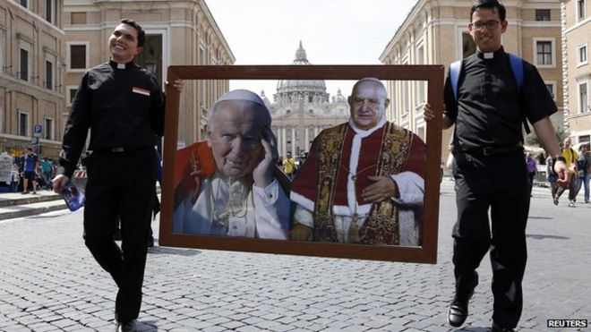 Двое священников гуляют с изображениями папы Иоанна Павла II (слева) и папы Иоанна XXIII на площади Святого Петра в Риме 25 апреля 2014 года.