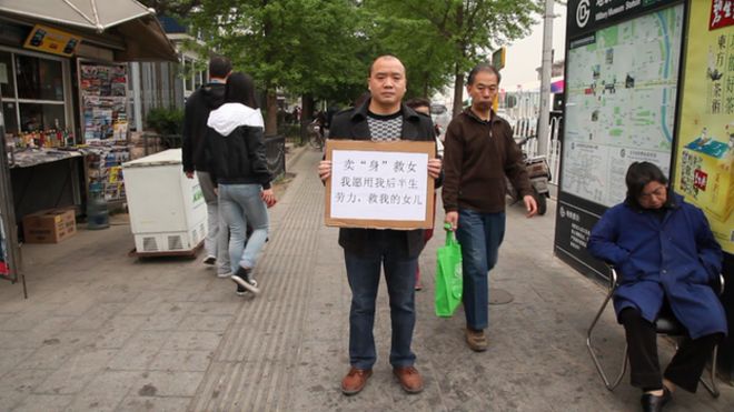 Чэн Банцзянь стоит на улице в Пекине и держит плакат с надписью: «Я буду работать всю оставшуюся жизнь для любого, кто финансирует медицинское обслуживание моей дочери».