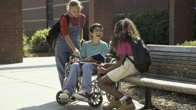 Мальчик в инвалидной коляске с двумя подругами, одна из них сидит на скамейке