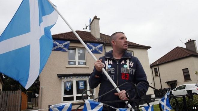 Шотландец машет шотландским флагом возле своего дома