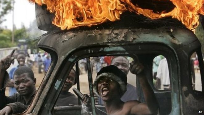 Кенийский мужчина сидит в кабине разрушенного грузовика, используемого в качестве временного блокпоста, в то время как шина горит на крыше, поскольку он и другие применяют контрольно-пропускной пункт во время насилия после выборов в Кисуму, Кения - вторник, 29 января 2008 г.