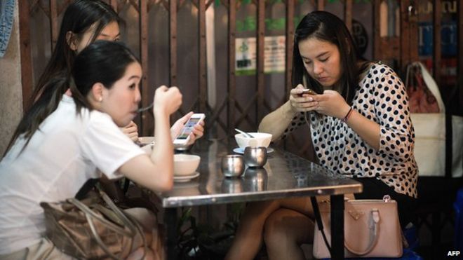 Группа молодых женщин, которые едят пищу, используя свои смартфоны