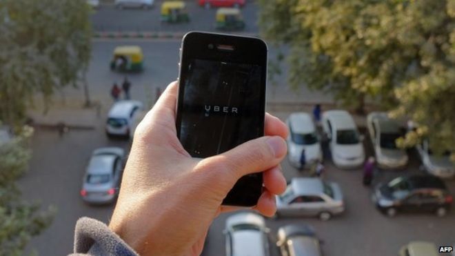Приложение Uber для смартфонов, которое используется для бронирования такси с помощью этой услуги, изображено над парковкой, когда авто-рикши (на заднем плане) проезжают по дороге в столице Индии Нью-Дели 7 декабря 2014 года
