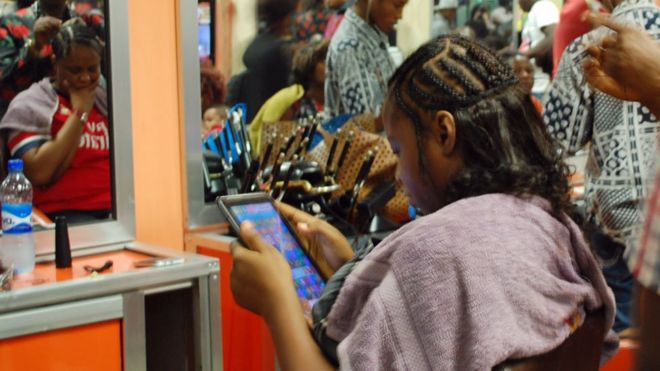 Клиент читает таблетку, когда ей делают прическу в парикмахерской Bruno's Place в торговом центре Ikeja Mall в Лагосе, Нигерия