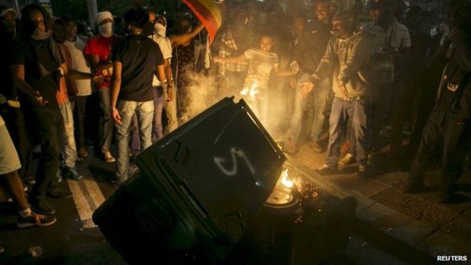 Протестующие, в основном израильские евреи эфиопского происхождения, стоят рядом с мусорным баком, который они подожгли на площади Рабина в Тель-Авиве 3 мая 2015 года.