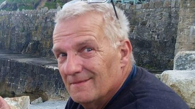 Melville Durrant - man in 60s, white/blond hair, glasses resting on top of head, partial smile, wearing black fleece, only face and shoulders in shot background of stone wall