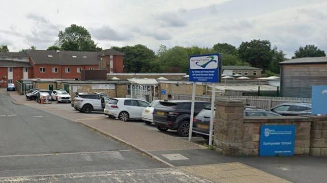 The entrance to the car park outside Springwater School in Starbeck. A blue sign on the right reads "Springwater School". Several cars are parked outside the sand-coloured, two-storey building.