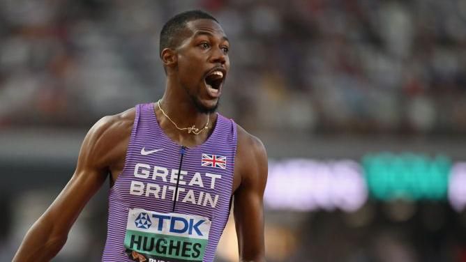 Team GB sprinter Zharnel Hughes on the celebrating on the track at the World Athletics Championships 2023.  He is wearing a purple and blue team GB track and field jersery.  He is in a running position with his mouth open.  He has a happy expression 