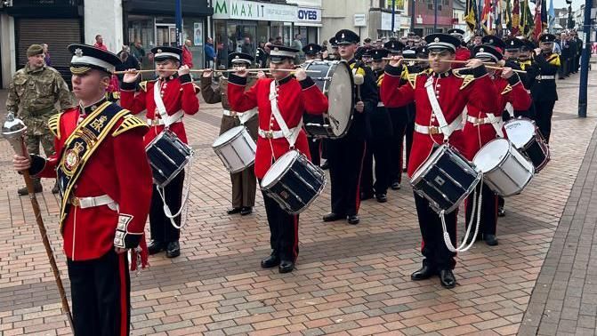 A parade of serving personnel drummers dressed in red and wearing gold Royal ER sashes being followed by marching veterans waving flags
