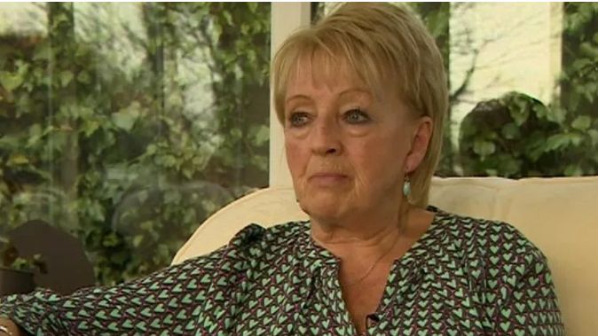 The victim's mother is sitting on a sofa with garden greenery visible through windows in the background. She is wearing a green dress and green drop earrings and her expression is sad