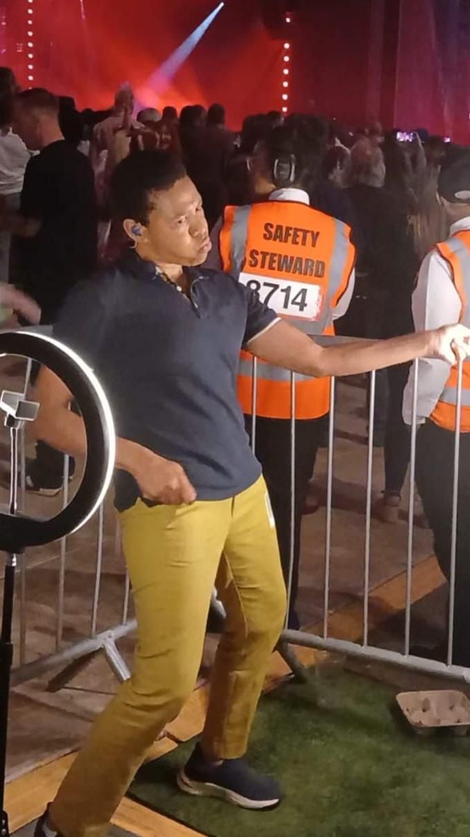 Ali Gordon wearing yellow trousers and a blue polo shirt with her hands out in sign language gestures and the stage and crowds behind her