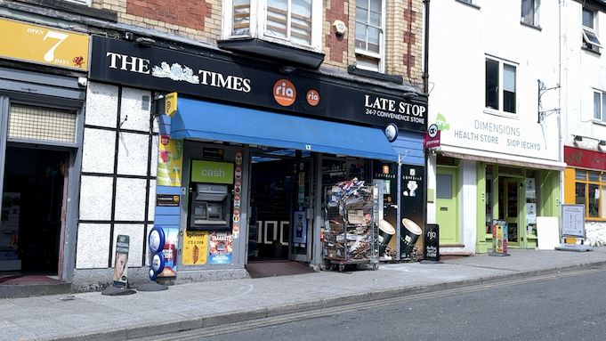 Exterior front of the Late Stop Shop, with a black hoarding showing The Times newspaper logo and the words Late Shop 24-7 Convenience Store, with a blue canopy below the sign, and a cash machine built into the shop front