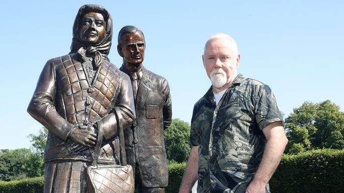 Anto Brennan posing with his sculpture. He is wearing a khaki sleeveless shirt and beige chinos. He is grey with short hair and a grey beard. 