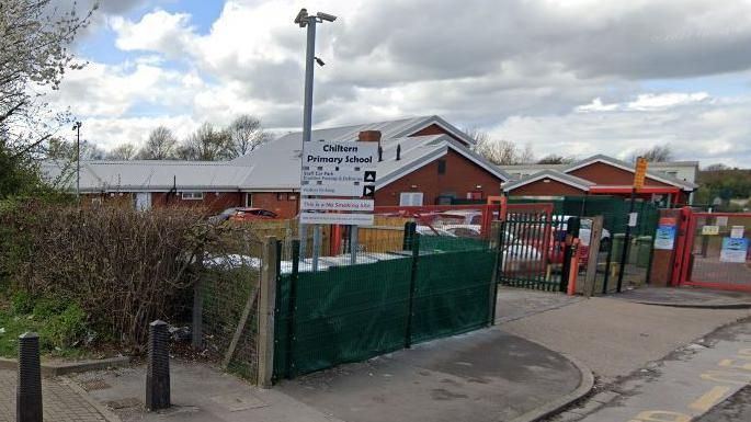 The front view of Chiltern Primary School. The building is low level with sloping grey roofs and has a red gate in at the front entrance.