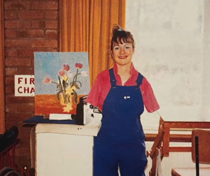 Alison Lapper, wearing a red top and blue dungarees, smiles in front of one of her paintings. The painting is of red and yellow flowers in a yellow vase. 