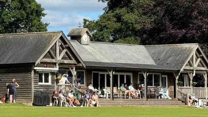 The clubhouse is shown with people sitting both on its decking and outside it as though they are watching a game.