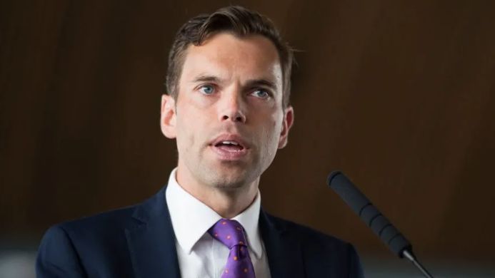 Ken Skates in white shirt, navy jacket and purple polka-dot tie, standing behind a mic