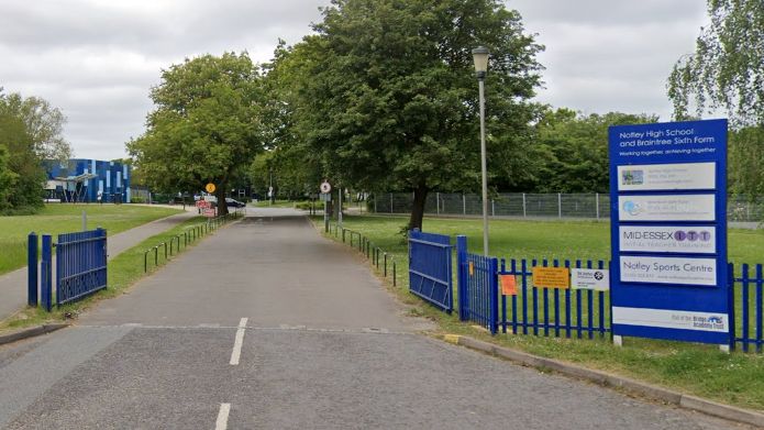 The entrance driveway to Notley High School & Braintree Sixth Form - a long road stretches towards the school premises and there is a blue school sign at the gate.