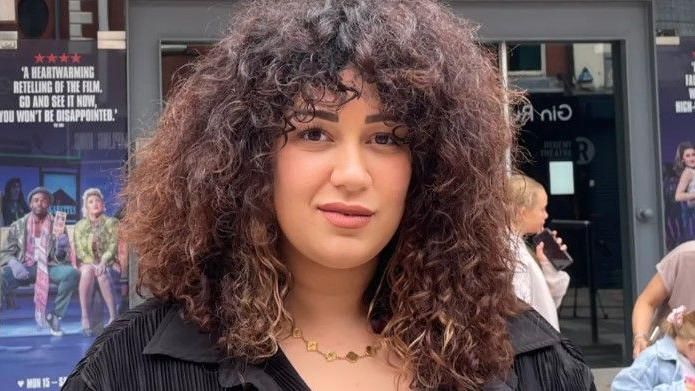 A woman with curly hair wearing a black jacket is stood in front of a theatre post with children stood in the background, one of whom is holding a mobile phone