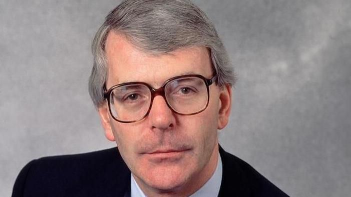 John Major wearing a suit and brown glasses, smiling directly at the camera