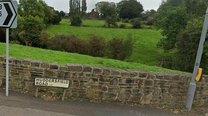 A road sign for Huddersfield Road in Brighouse at the side of the road against a brick wall with fields and trees behind